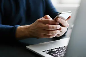 A person sitting at their computer with a phone in their hands